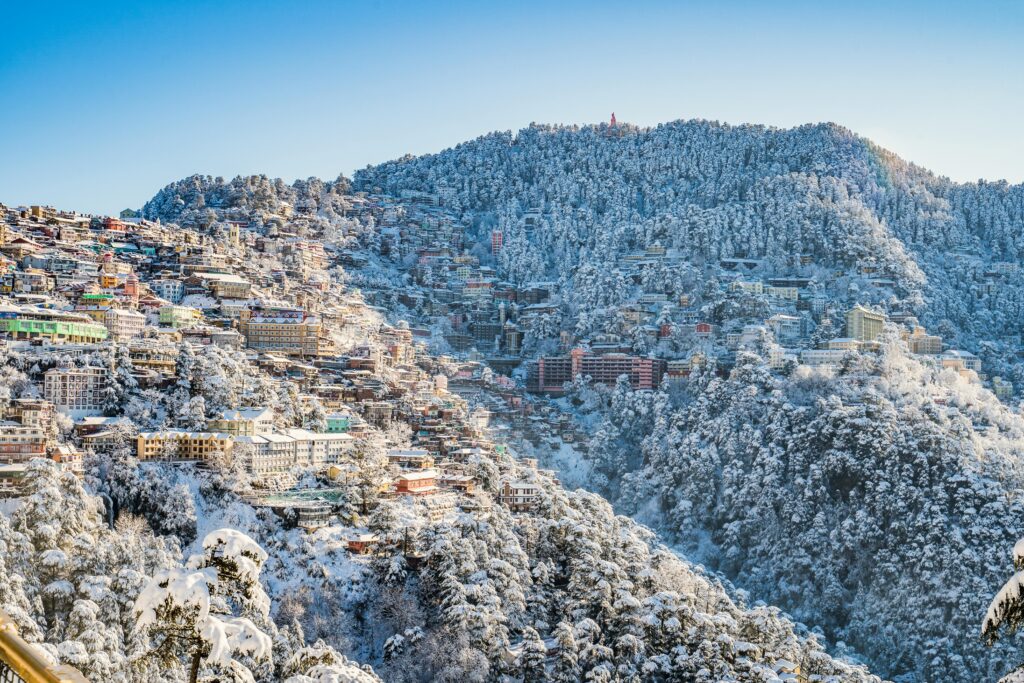 A scenic view of Shimla in winter, with snow-covered buildings and forests under a clear blue sky.